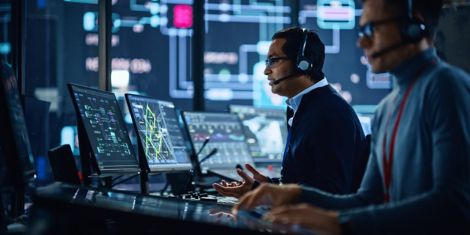 Portrait of Professional IT Technical Support Specialist Working on Computer in Monitoring Control Room with Digital Screens. Employee Wears Headphones with Mic and Talking on a Call.