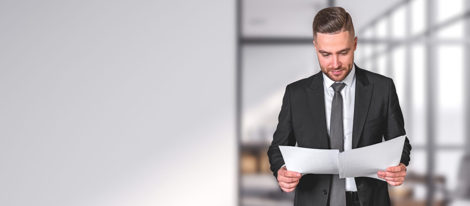 Businessman reading business documents in hands. Concentrated man portrait in black formal suit, studying financial report. Copy space empty white office wall. Concept of contract