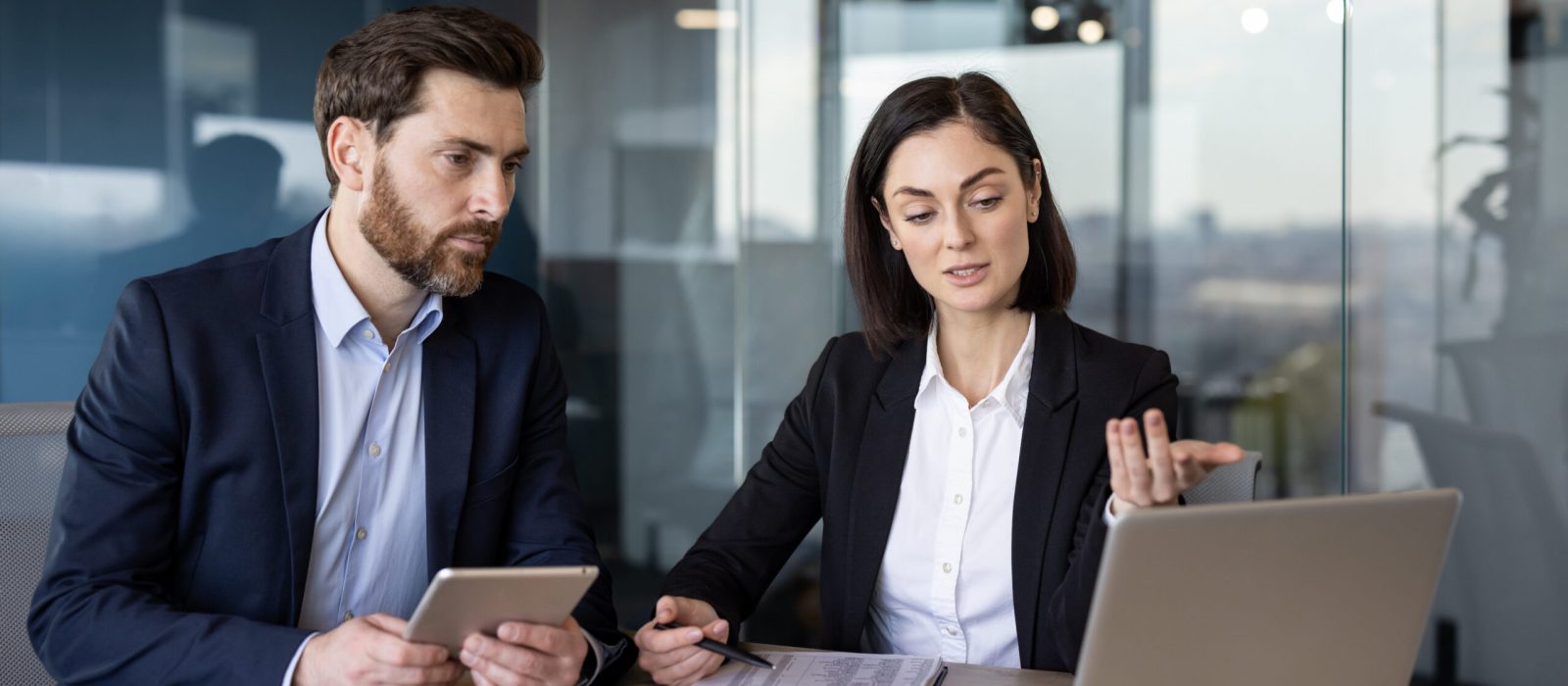 Successful business partners sitting by table with digital devices and looking at pc screen attentively. Caucasian woman and man in formalwear discussing and analyzing statistics of sale season.