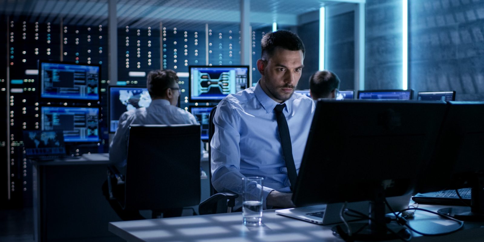 Close-up of Technical Engineer Working on His Computer With Multiple Displays in Monitoring Room. In the Background His Colleagues with Graphics and Various Data on Their Monitors.