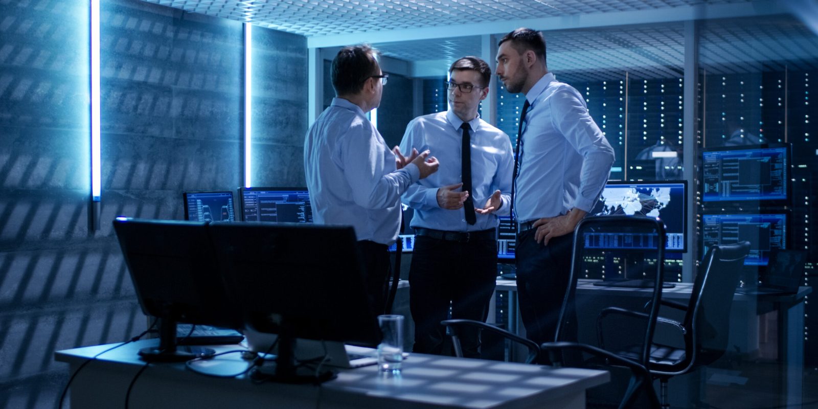 Team of Technical Moderators Have Active Discussion in Monitoring Room. System Control Room is full of Working Displays Showing Various Data and Has Servers Racks.