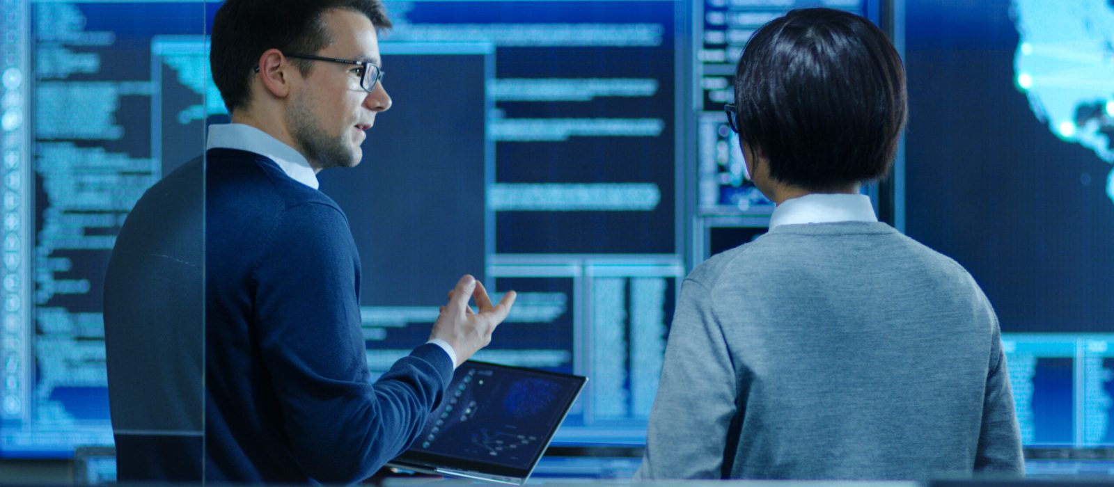 In the System Control Room IT Specialist and Project Engineer Have Discussion while Holding Laptop, they're surrounded by Multiple Monitors with Graphics. They Work in a Data Center on Data Mining, AI and Neural Networking.